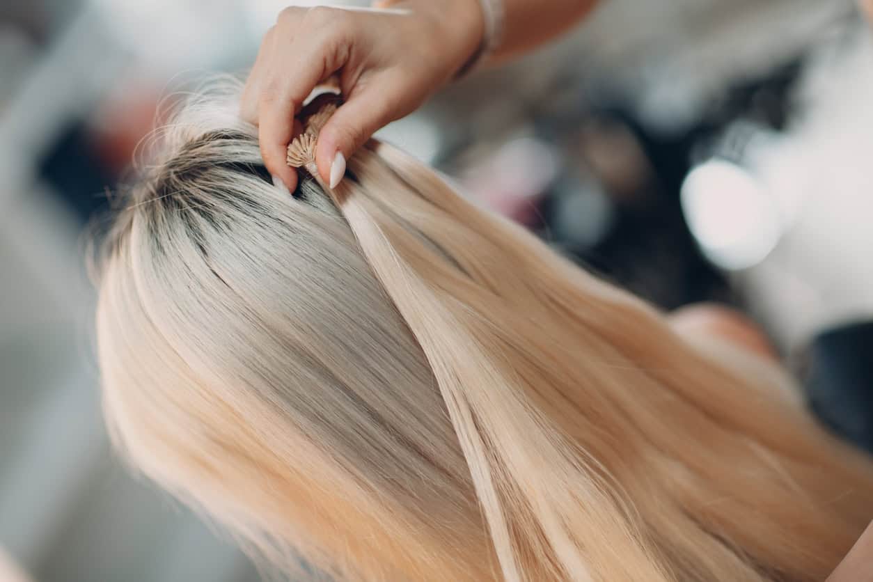 Hairdresser female making hair extensions to young woman with blonde hair in beauty salon. Professional hair extension.